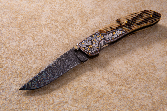 The "Protege" Fine Folder, fitted with a 3 3/8" Damascus blade, FLuted Mammoth ivory scales, engraved bolstes by Alice Cartar, and fileworked, jeweled and anodized backspacer and liners.